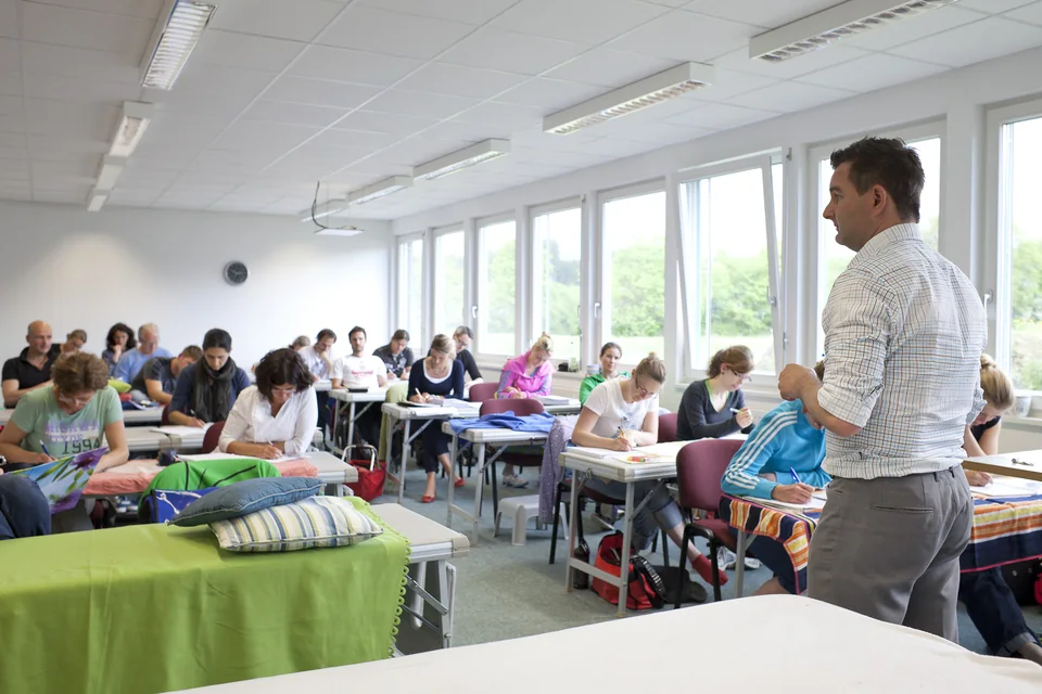 Kursraum während einer Kurswoche: Studierende an den Tischen, der Dozent geht durch die Reihen