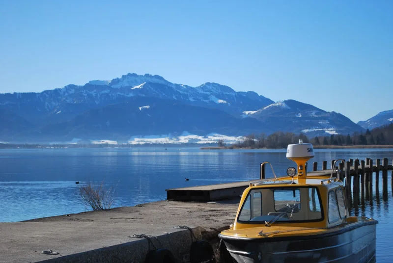 Boote am Steg, dahinter der Chiemsee und die verschneiten Alpen